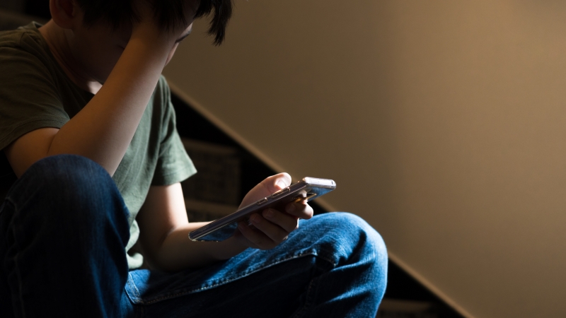 Ein Kind sitzt auf der Treppe, in der einen Hand hält er ein Smartphone, mit der anderen stützt er seinen Kopf