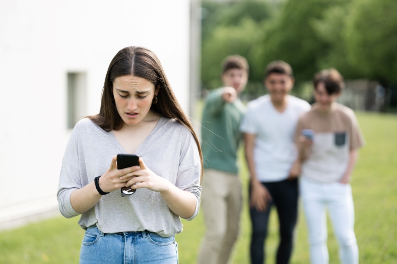 Im Vordergrund steht ein Mädchen, das erschrocken auf ihr Smartphone blickt, im Hintergrund stehen lachende Kinder